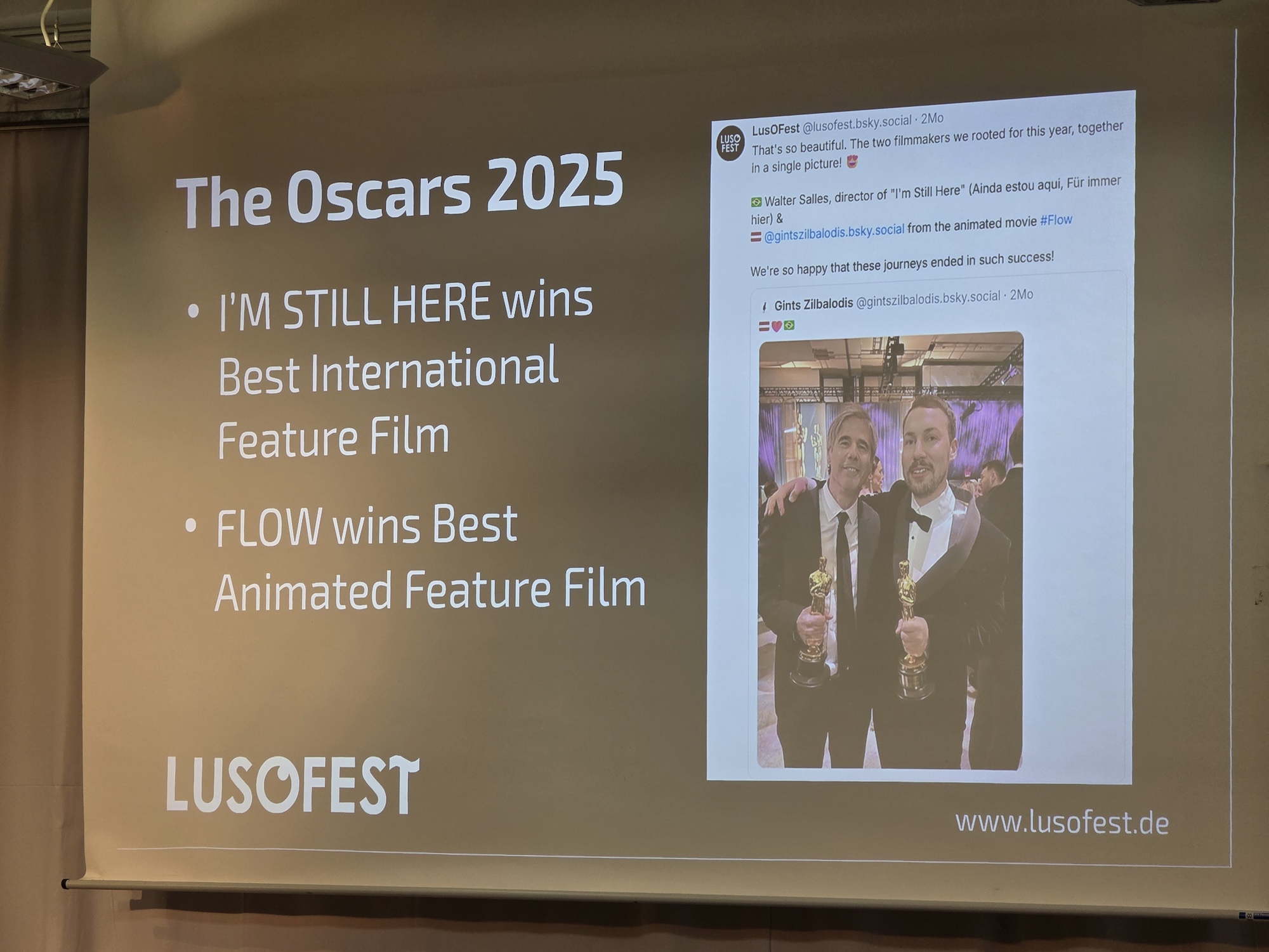 A photo of a slide with the sayings 'The Oscars 2025', 'I'm Still Here' wins Best International Feature Film, 'Flow' wins Best Animated Feature Film, and a picture of directors Walter Salles and Gints Zilbalodis hugging and holding their awards. Lusofest.